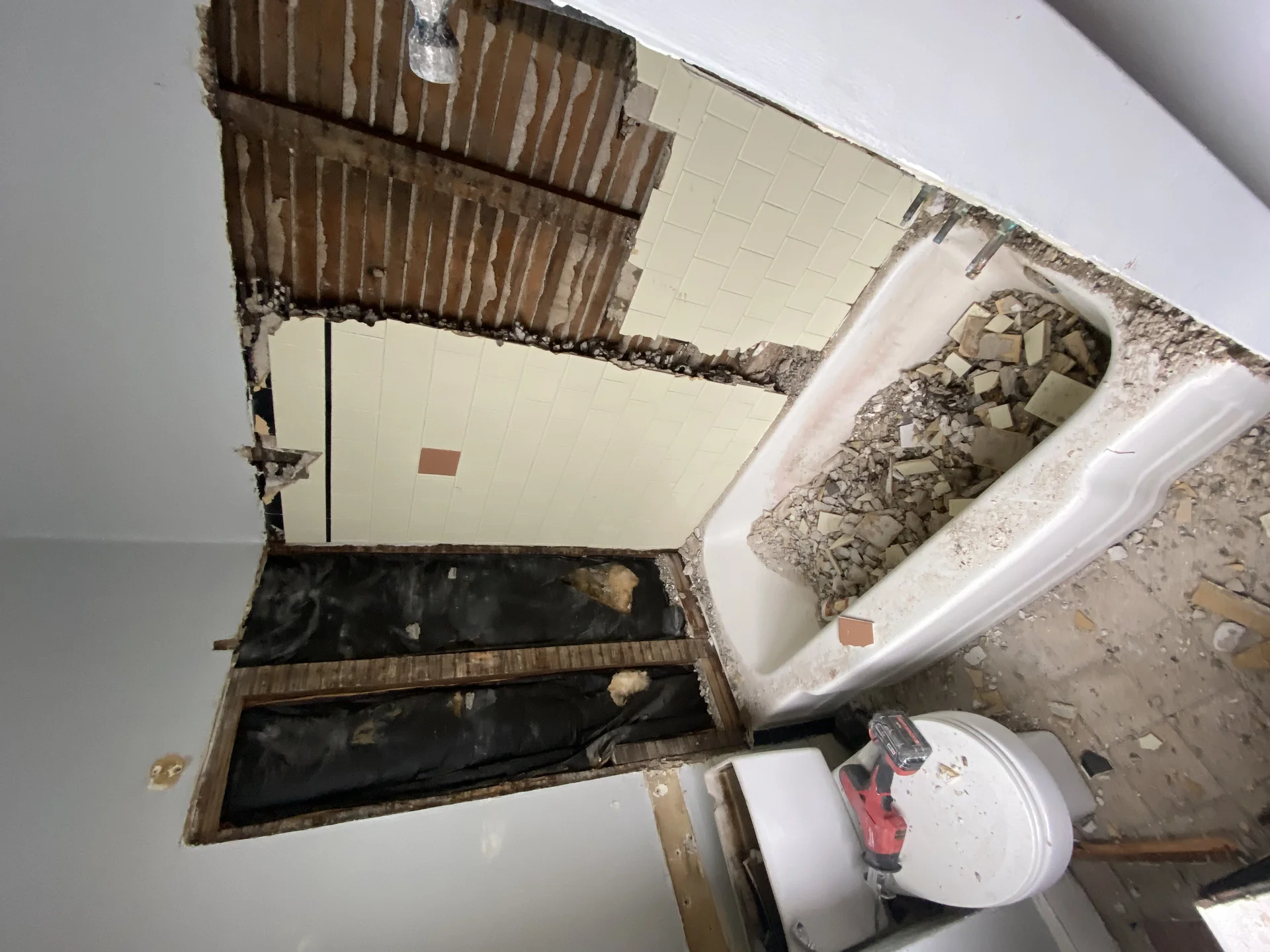 Bathroom undergoing demolition with old tiles removed, exposing lath and plaster walls, and a tub full of debris. by SF Const