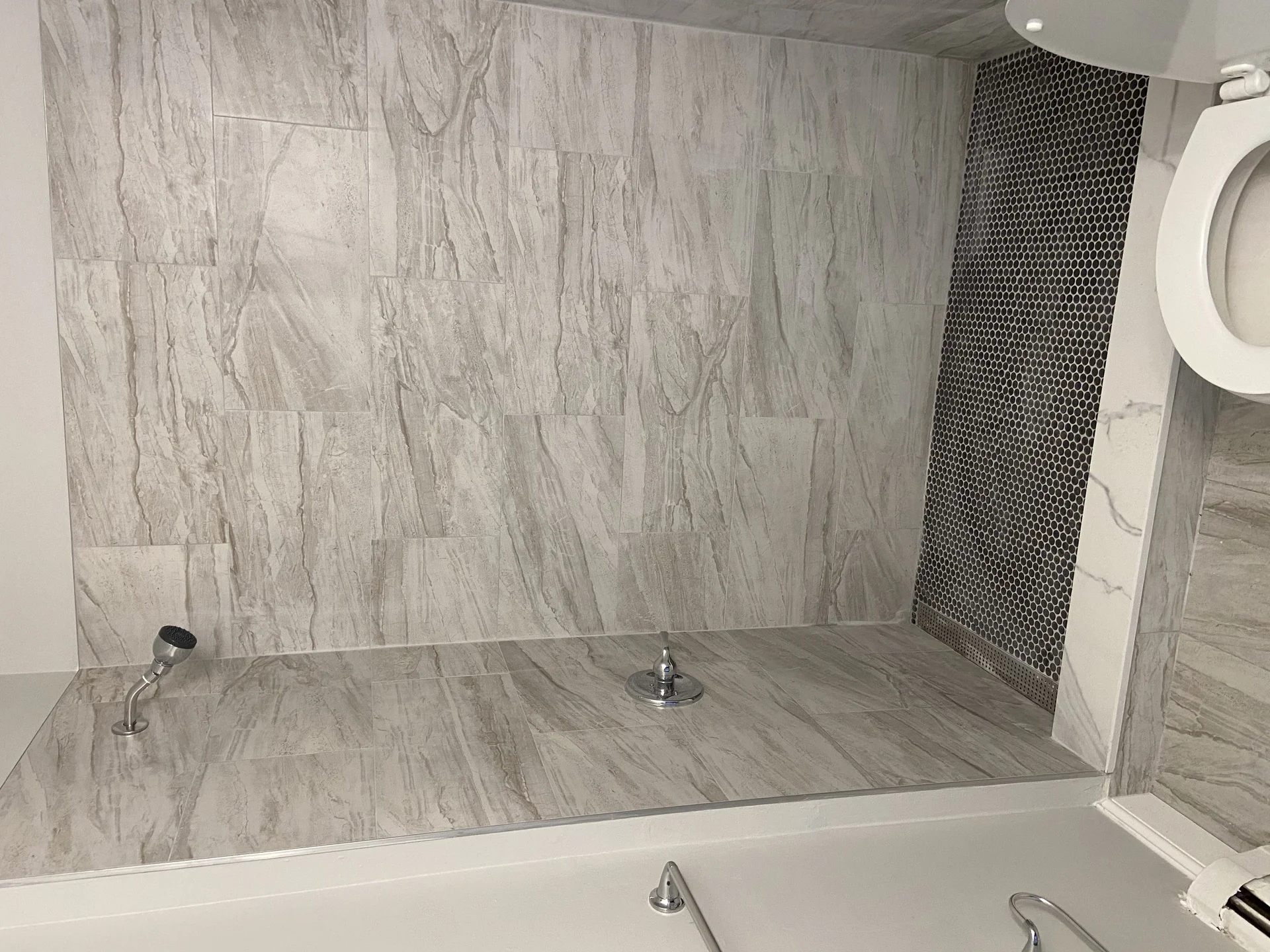 Modern shower with light grey marble-look tiles, dark hexagonal floor tiles, and chrome fixtures. by SF Construction in Hudso