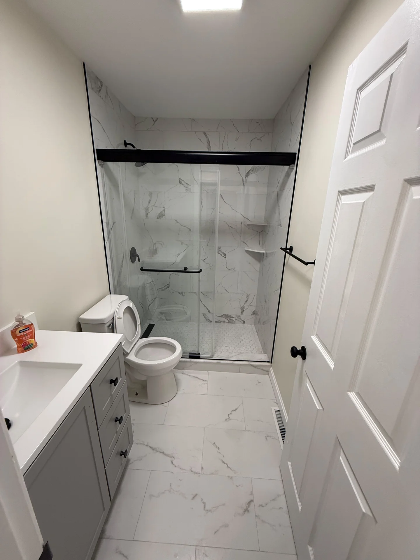 Bright, modern bathroom with white marble-look tiles, grey vanity, glass shower, and black fixtures. by SF Construction in Hu