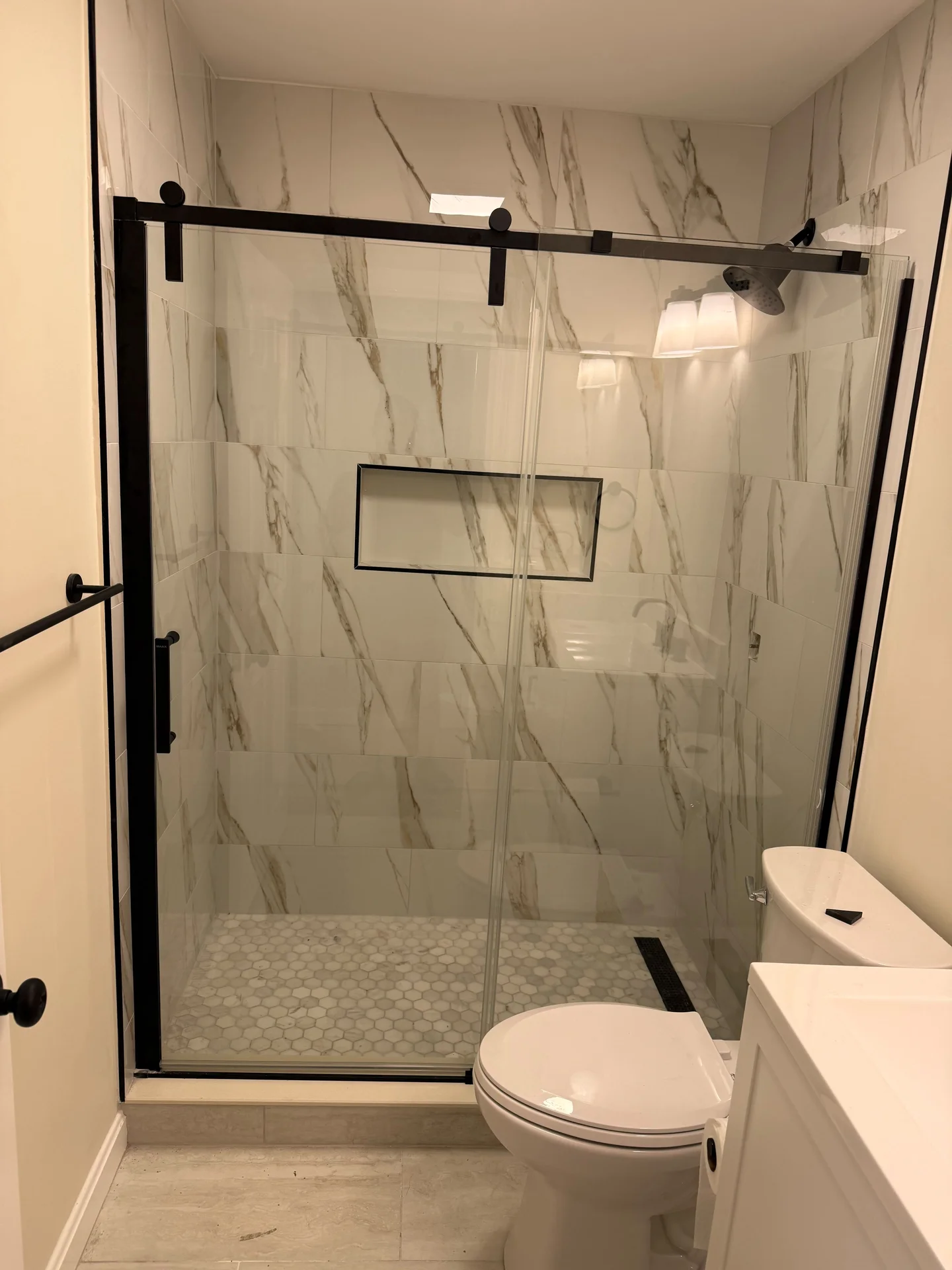 Modern bathroom with white marble-look shower tiles, hexagon shower floor, and black-framed glass door. by SF Construction in