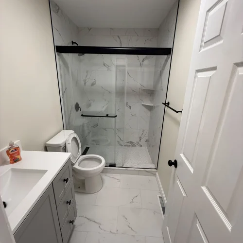 Bright, modern bathroom with white marble-look tiles, grey vanity, glass shower, and black fixtures. by SF Construction in Hu