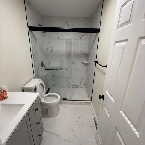 Bright, modern bathroom with white marble-look tiles, glass shower, grey vanity, and black fixtures. by SF Construction in No